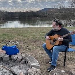 Chapman playing an acoustic guitar beside a lake